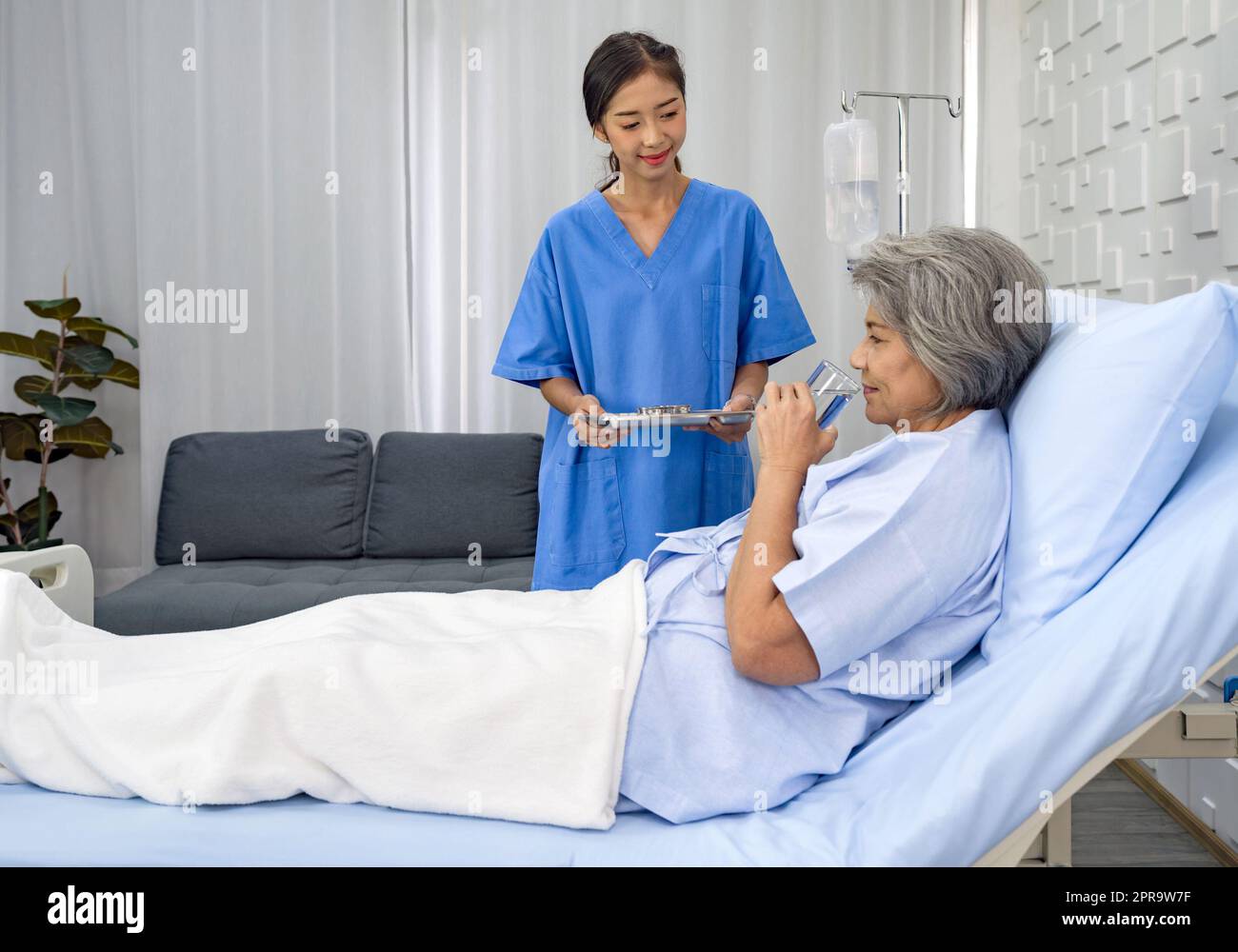 Un patient asiatique âgé à poil blanc reçoit un médicament et un verre d'eau de la part d'une infirmière. Une femme âgée étant traitée à l'hôpital, allongée sur le lit. Banque D'Images