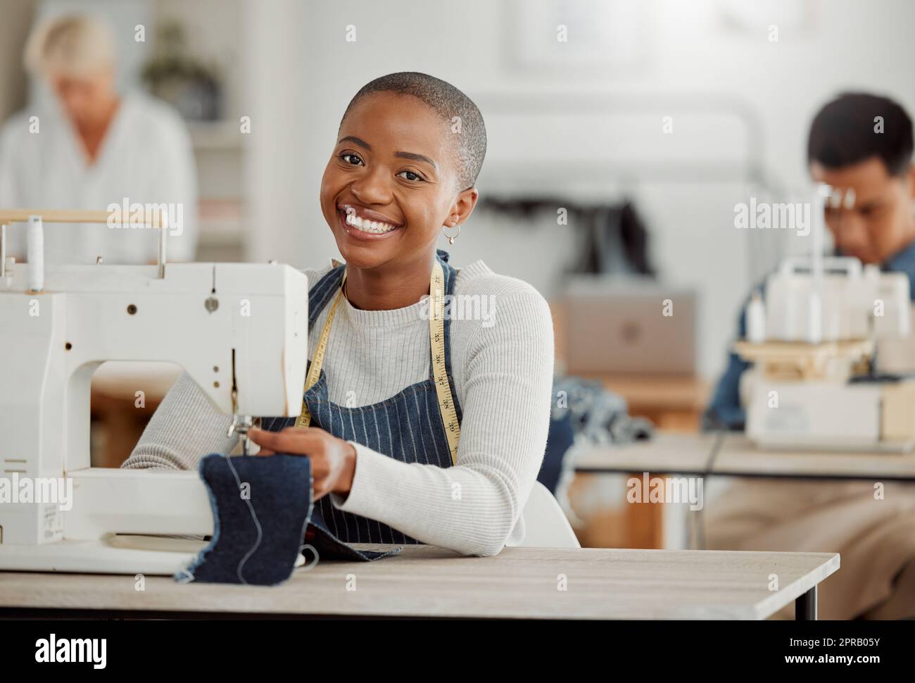 Joyeux, créatif étudiant de mode noir ou designer assis et souriant en classe par une machine à coudre travaillant sur des vêtements. Portrait isolé d'un magnifique tailleur afro-américain dans une usine ou un studio. Banque D'Images