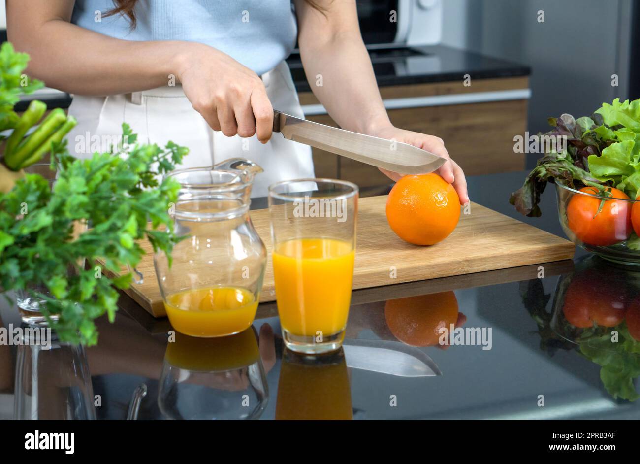 Gros plan main tenant le couteau coupant des fruits orange sur une planche à découper en bois. Un bol contenant un mélange de jus de fruits et un bol en verre contenant une variété de fruits et légumes est placé sur le comptoir de la cuisine. Banque D'Images