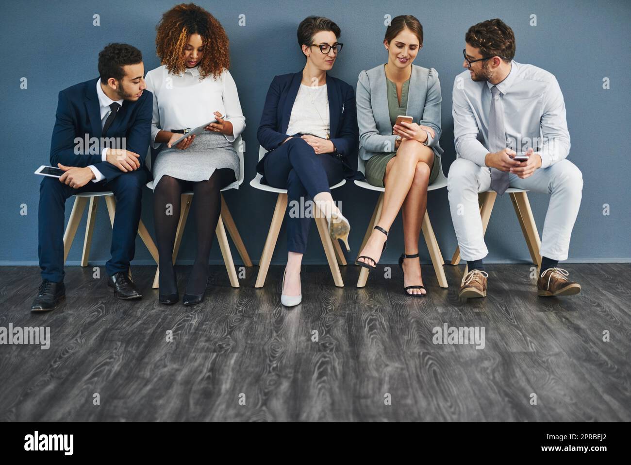 Groupe de gens d'affaires en réseau sur des téléphones, des tablettes numériques et des médias sociaux, en attente dans la file d'attente. Une équipe diversifiée de collègues assis dans la file d'attente, parlant et discutant contre le mur bleu d'espace de copie. Banque D'Images