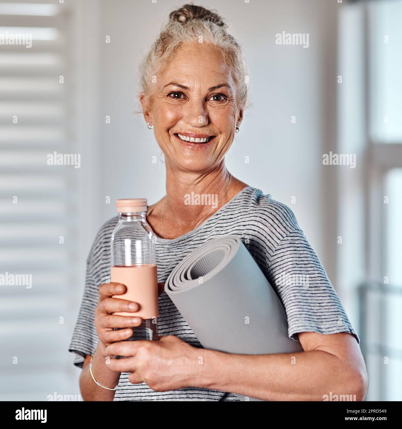 J'ai tout ce dont j'ai besoin pour commencer mon entraînement. Portrait d'une femme mûre et gaie tenant une bouteille d'eau et un tapis de yoga prêt à commencer sa séance matinale de yoga à l'intérieur d'un studio. Banque D'Images