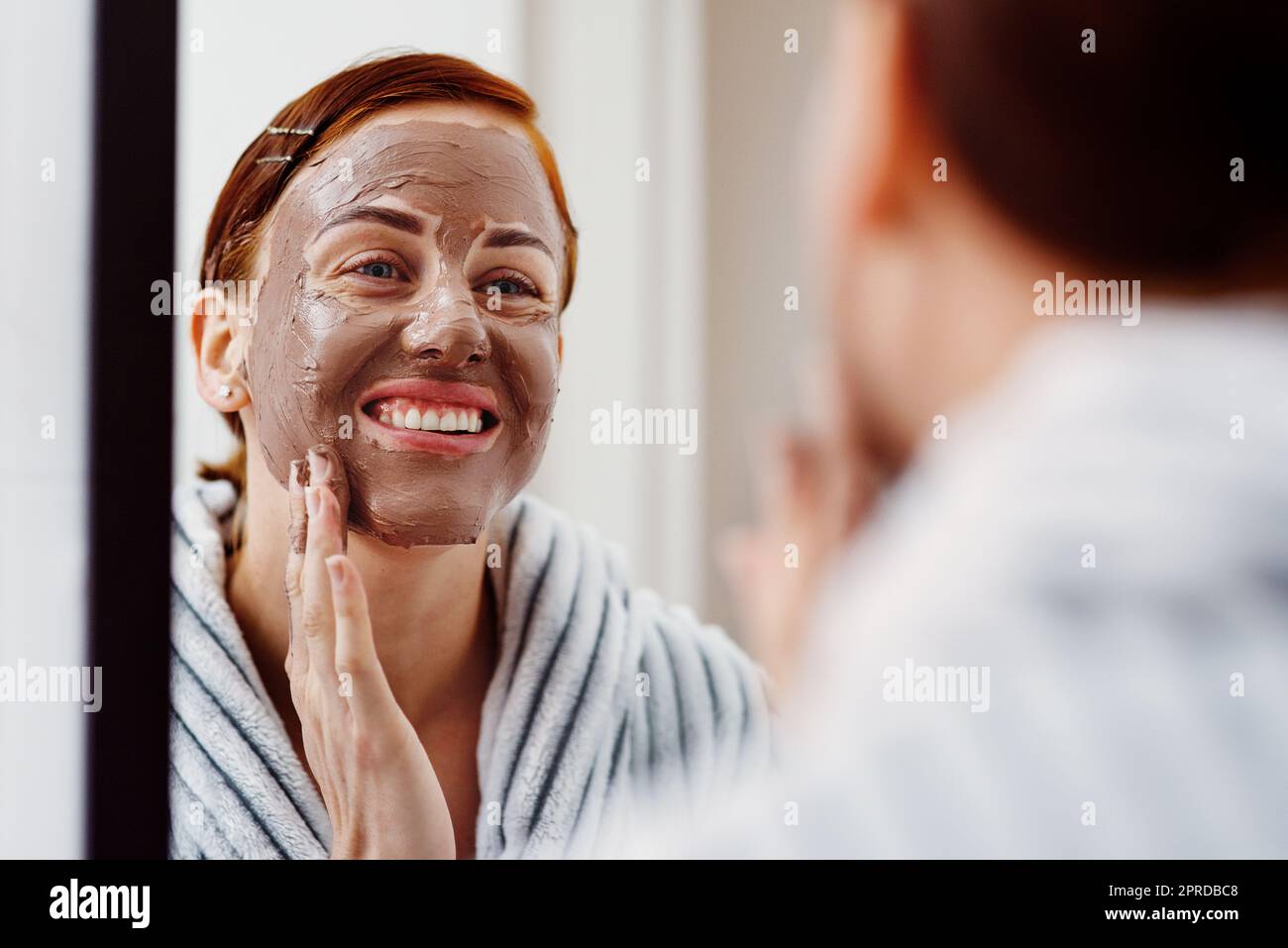 Masqué avec beauté. Une jeune femme attrayante appliquant un masque de boue sur son visage tout en se tenant devant le miroir dans sa salle de bains à la maison. Banque D'Images