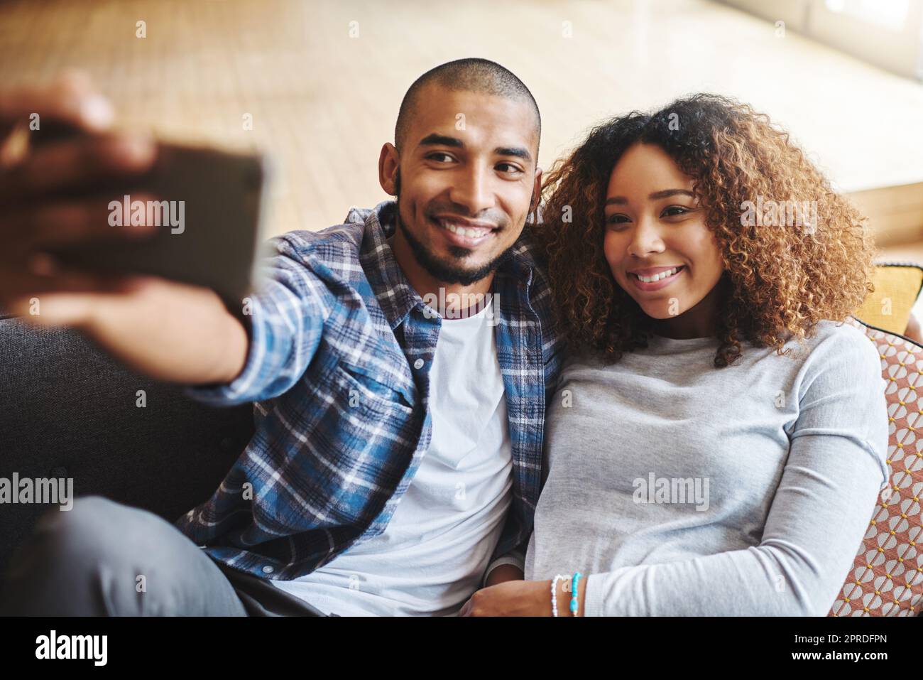 Souriez babe. Heureux, romantique et amoureux couple de prendre un selfie ensemble sur le téléphone assis sur le canapé. Jeunes conjoints souriants se tenant dans le téléphone pour prendre une photo dans le salon. Banque D'Images