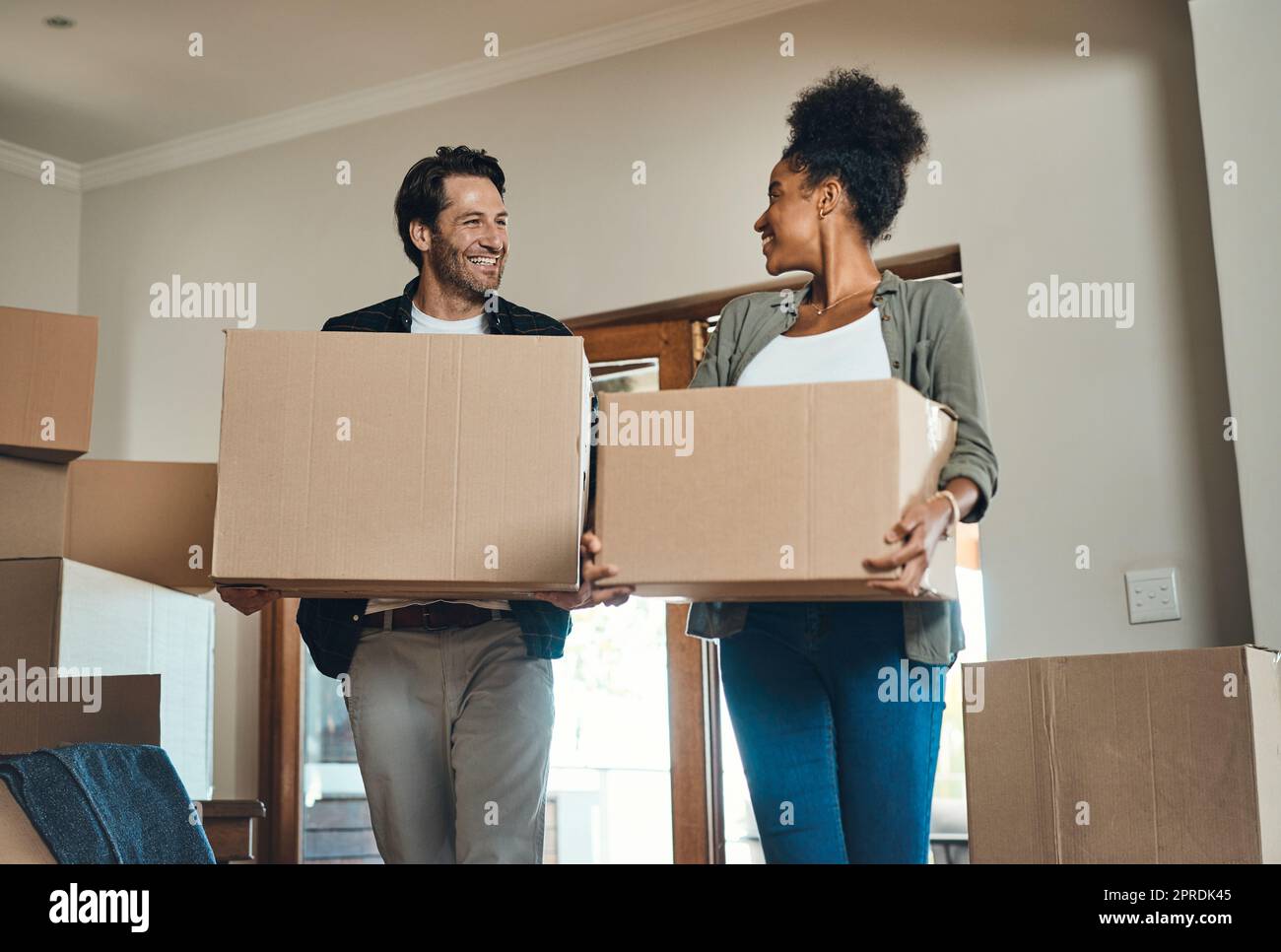 Un couple heureux ou de nouveaux propriétaires de maison se déplaçant ensemble, portant des boîtes de meubles ou d'effets personnels et de la propriété. Des partenaires amoureux, interraciaux après l'achat de biens immobiliers, souriant les uns les autres. Banque D'Images