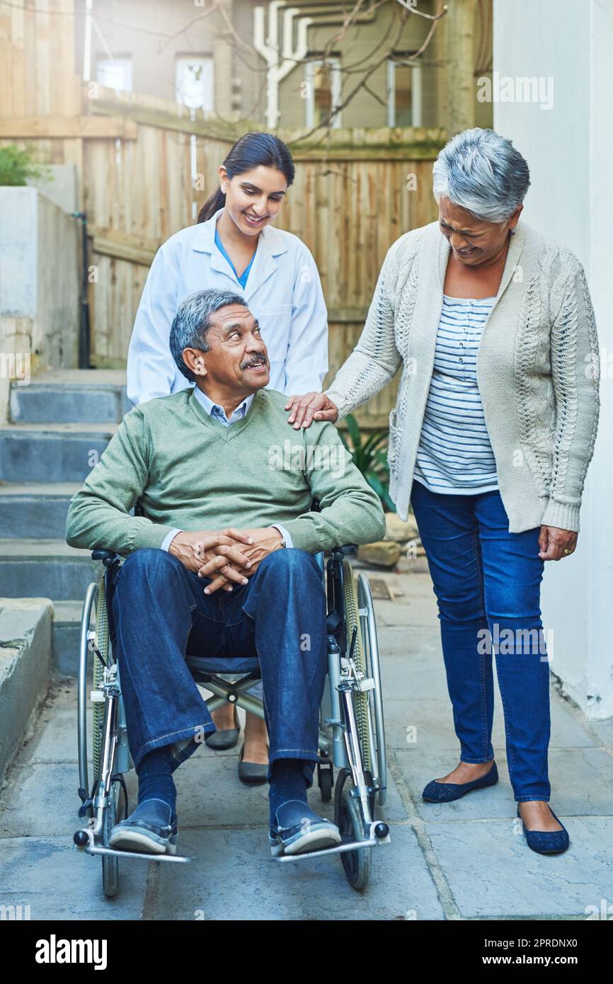 Où que vous alliez, nous allons. Un homme âgé gai assis dans un fauteuil roulant tout en étant soutenu par sa femme et une infirmière féminine à la maison pendant la journée. Banque D'Images
