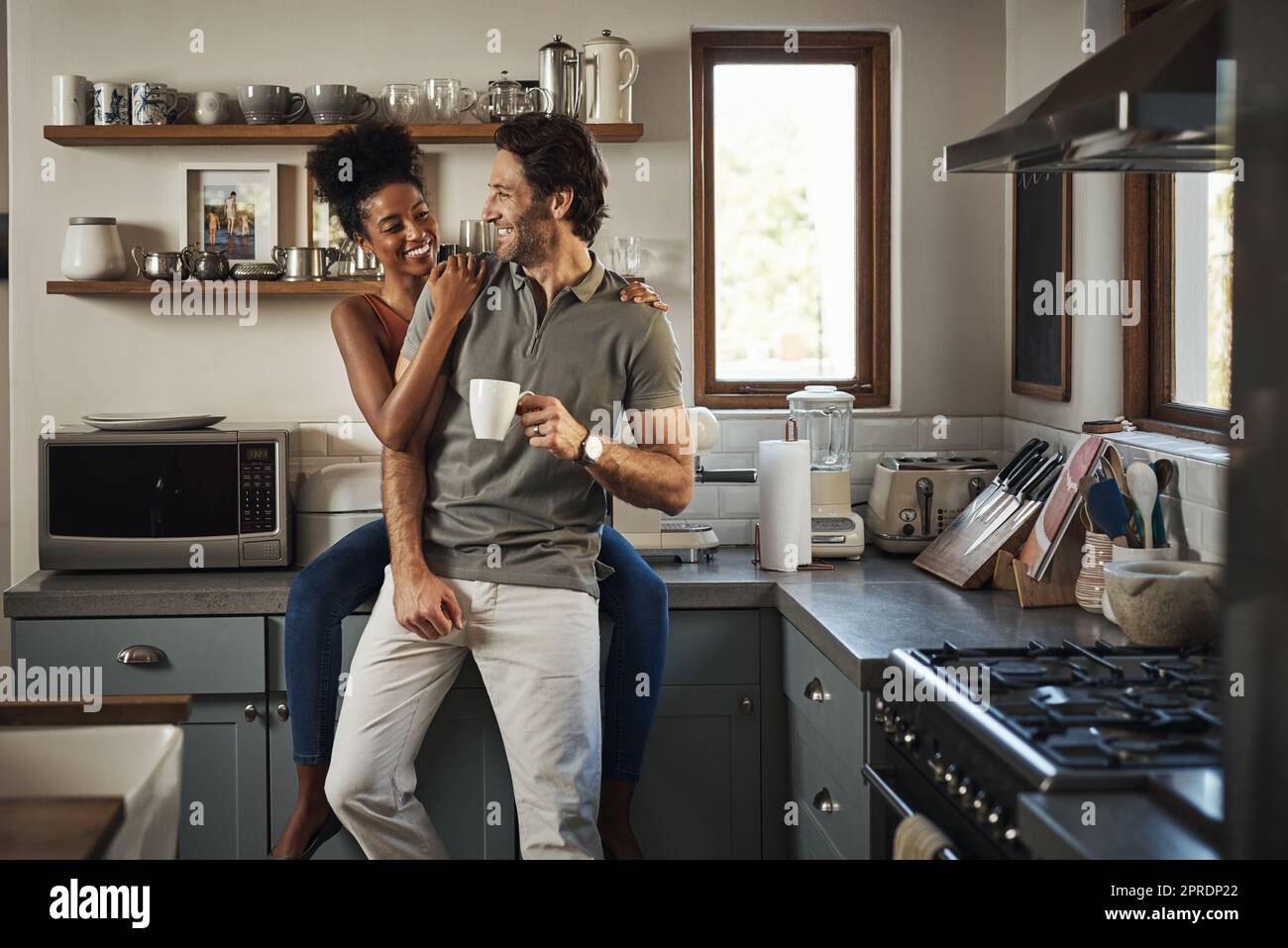 Heureux, dans l'amour et rire tandis qu'un couple interracial aime le café du matin et le collage tout en ayant une bonne communication dans une relation. Mari et femme parlant en se tenant dans la cuisine à la maison Banque D'Images