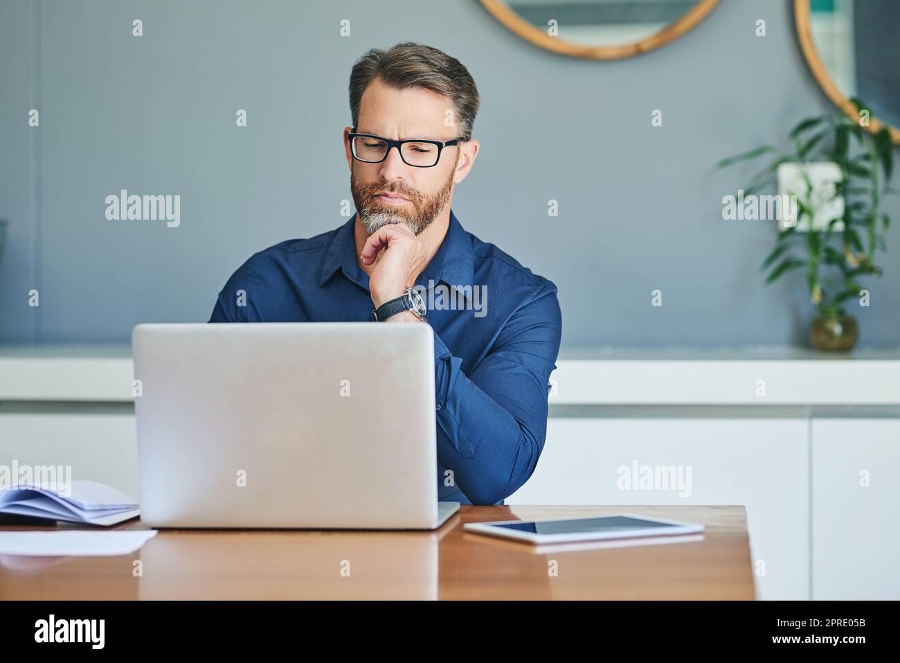 Faire du travail à la maison. Un homme concentré d'âge moyen qui travaille sur son ordinateur portable tout en contemplant à la maison pendant la journée. Banque D'Images