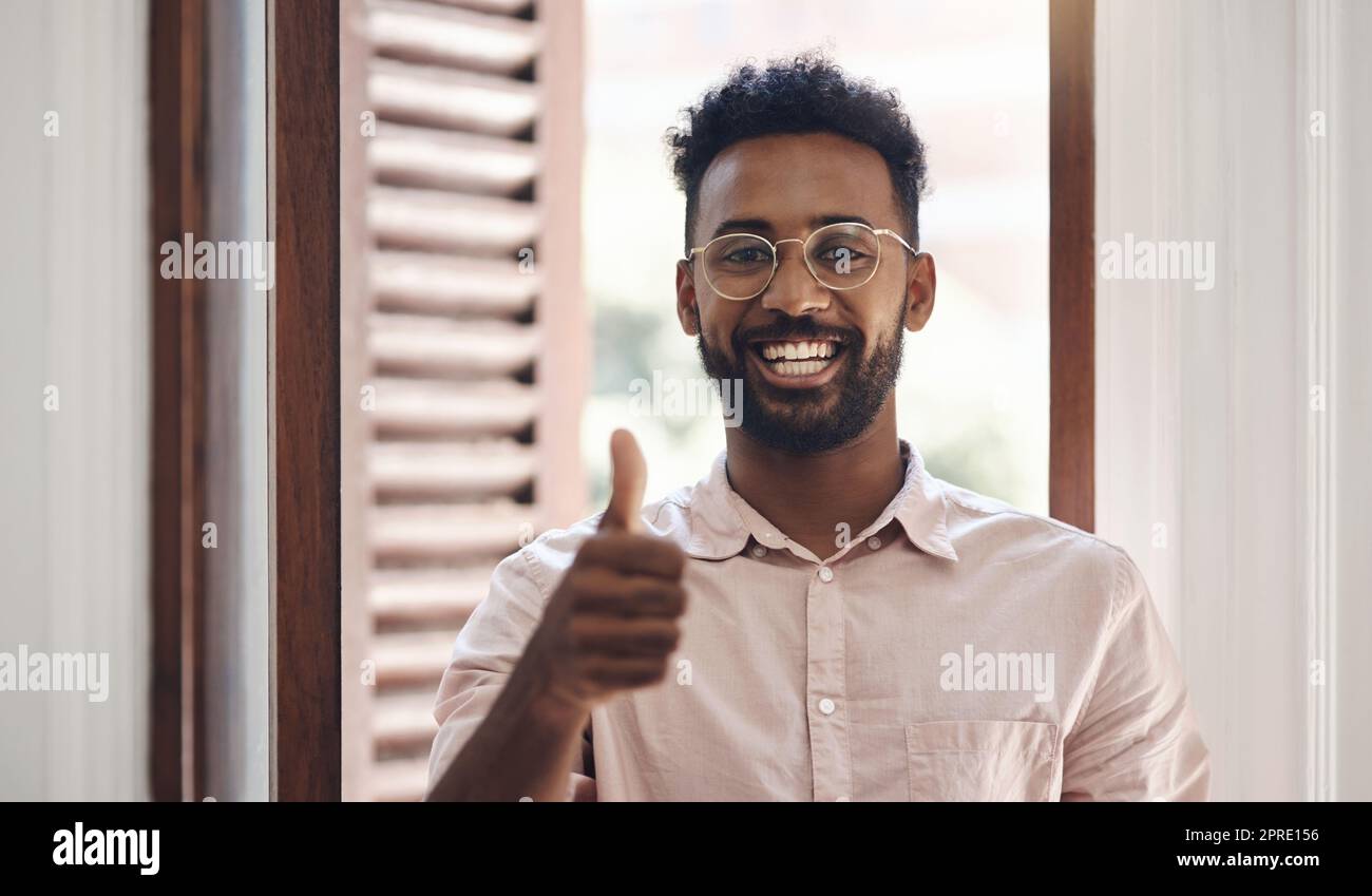 Pouces vers le haut, endossant et merci avec un jeune homme souriant, se sentant positif et donnant son approbation tout en se tenant à l'intérieur. Portrait d'un homme heureux portant des lunettes et donnant sa confiance ou son soutien Banque D'Images