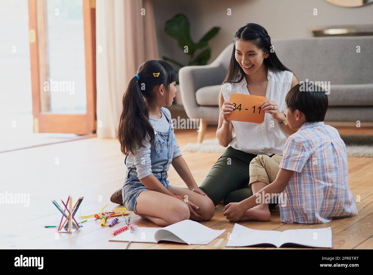 Pensez dur et soigneusement. Une mère gaie et ses deux enfants faisant leurs devoirs ensemble tout en étant assis sur le sol à la maison pendant la journée. Banque D'Images