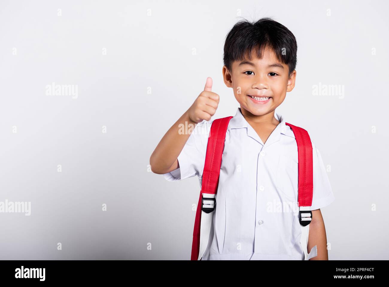 Asiatique adorable tout-petit souriant heureux portant étudiant thaïlandais uniforme pantalon rouge montrer pouce vers le haut de doigt Banque D'Images