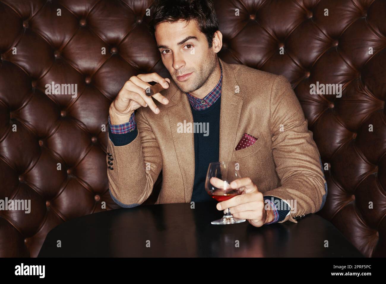 J'aime un bon cigare avec mon whisky. Portrait d'un beau jeune homme qui boit et fume un cigare dans un club Banque D'Images