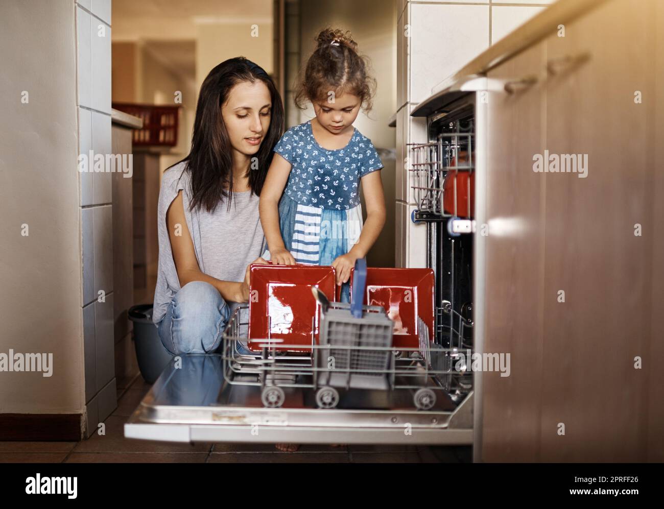 Élever une petite fille responsable. Une petite fille mignonne et sa mère chargeant le lave-vaisselle ensemble à la maison. Banque D'Images