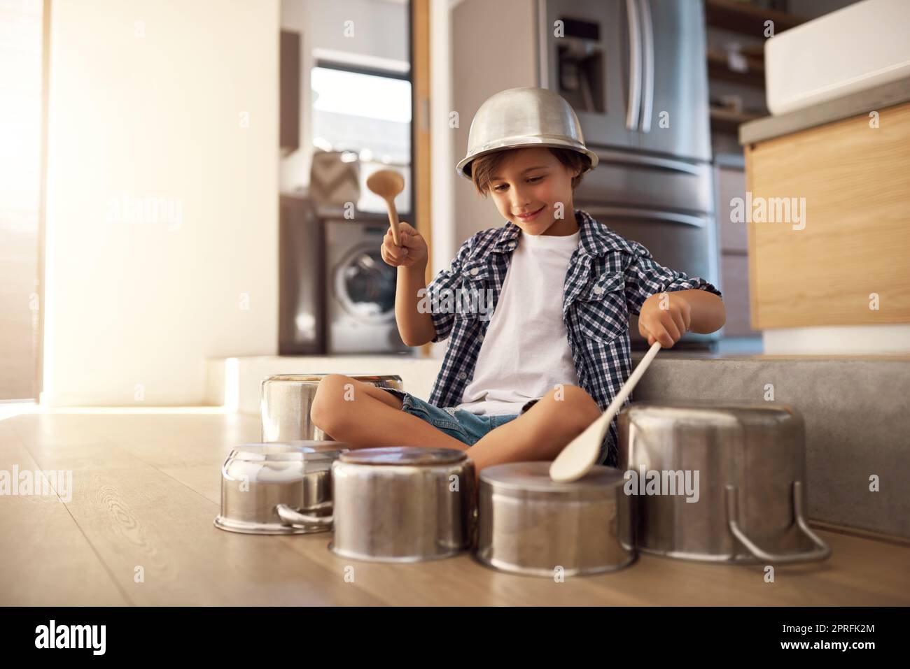 HES un peu bruyant. Un petit garçon heureux jouant des tambours avec des pots sur le sol de la cuisine tout en portant un bol sur sa tête. Banque D'Images