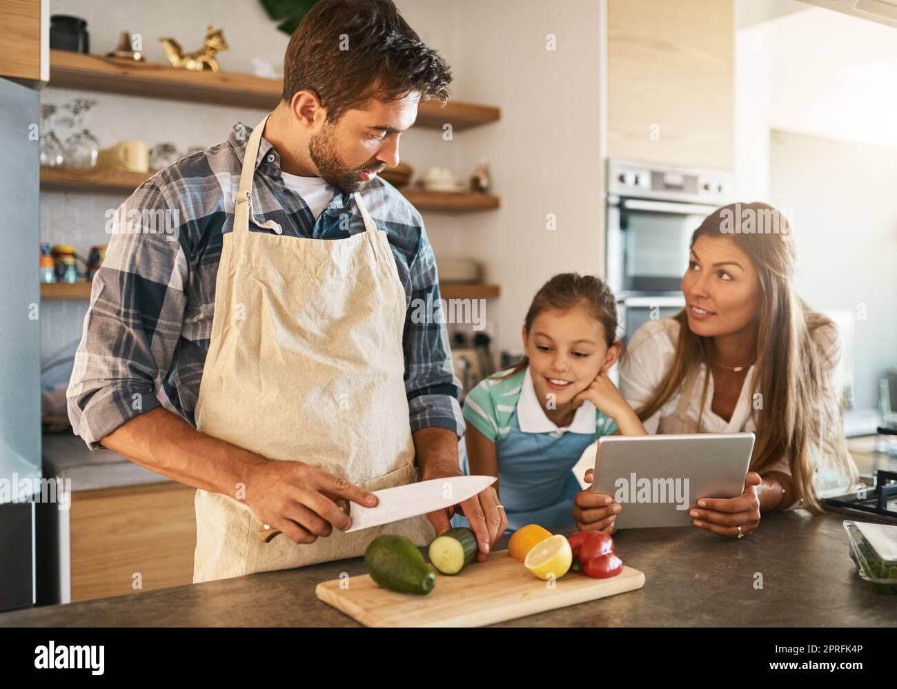 Les couteaux sont pour adultes seulement, d'accord. Deux parents heureux et leur jeune fille essayant une nouvelle recette dans la cuisine ensemble. Banque D'Images