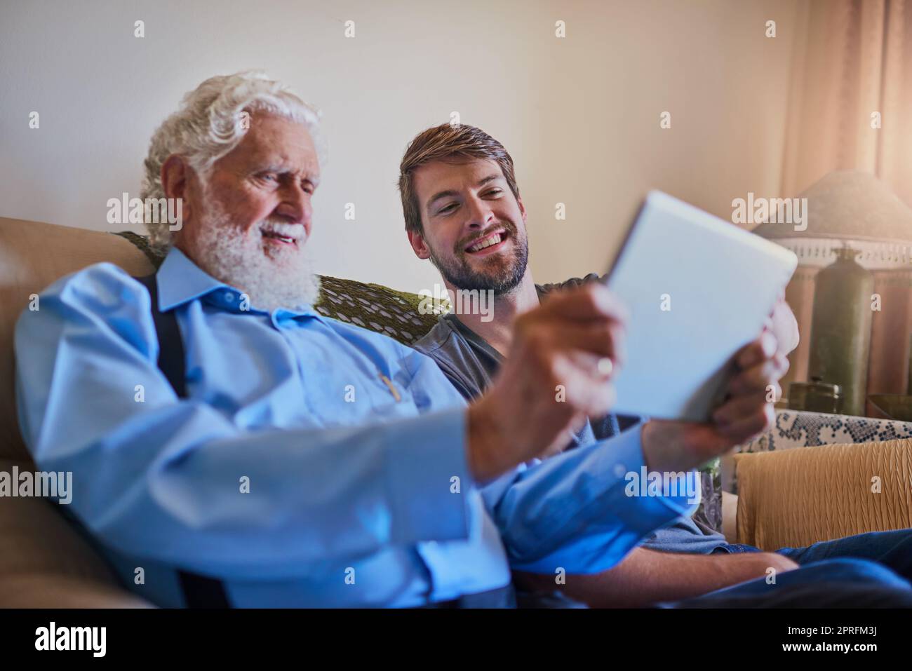 Laissez votre grand-père vous montrer une ou deux choses. Un jeune homme montrant à son grand-père âgé comment utiliser une tablette tout en étant assis sur le canapé à la maison. Banque D'Images