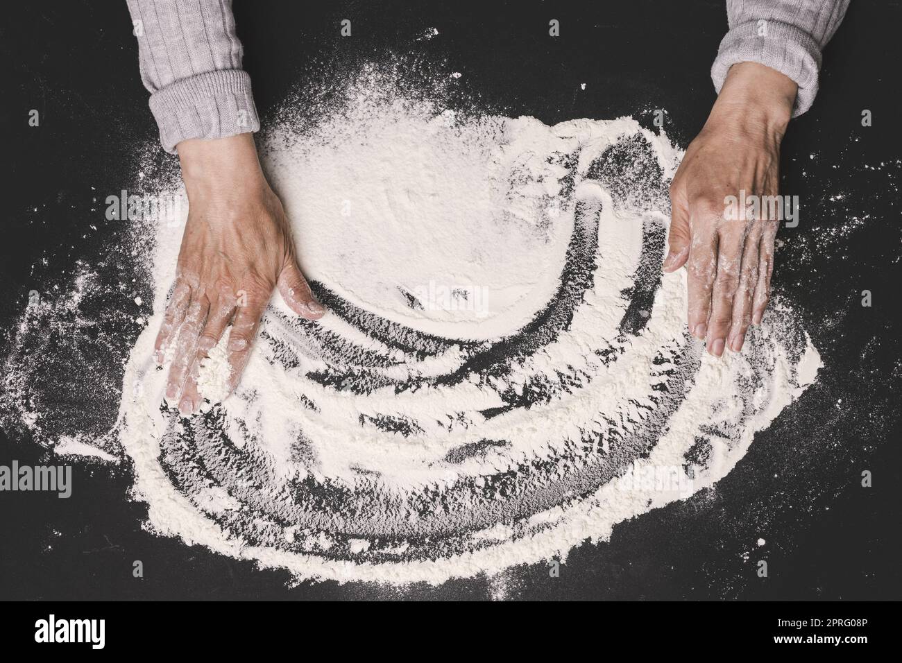 Farine de blé blanc tamisée sur une table noire et deux mains femelles, vue du dessus. Cuisiner à la maison Banque D'Images