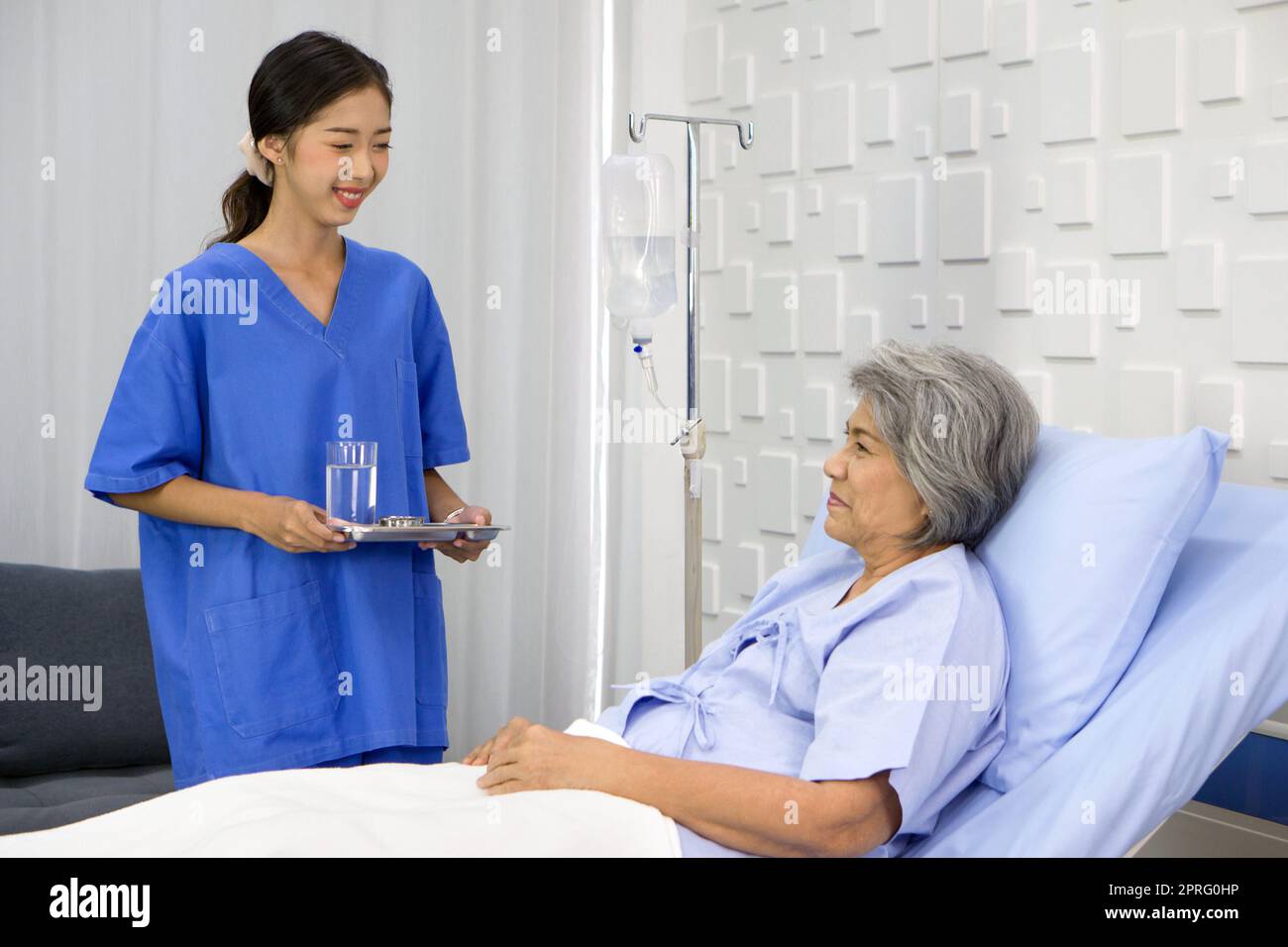 Une jeune infirmière asiatique en uniforme bleu apporte un médicament et un verre d'eau aux patients âgés à cheveux blancs. Une femme plus âgée étant traitée à l'hôpital, allongée sur le lit. Banque D'Images