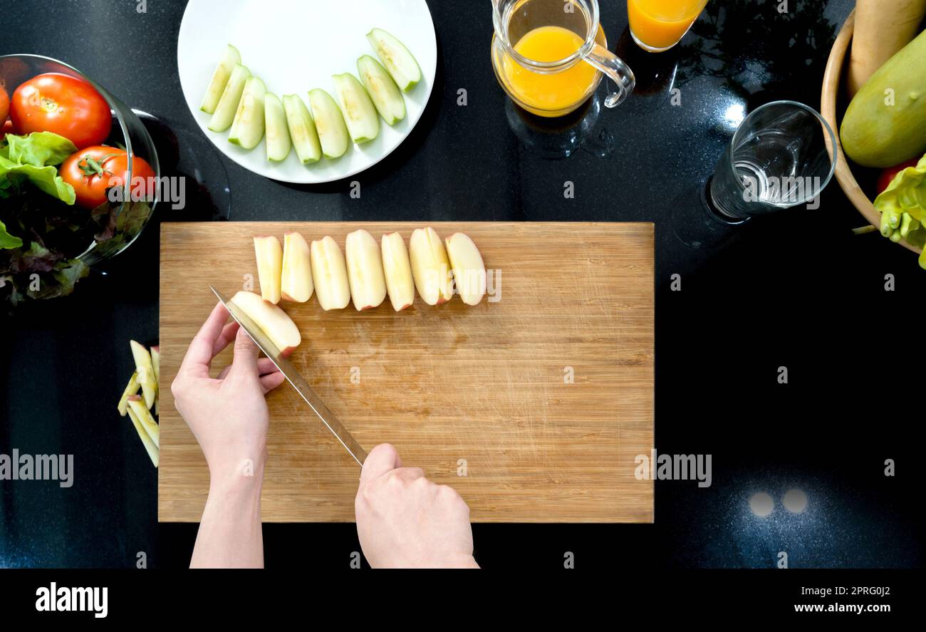 Gros plan main tenant le couteau coupant la pomme rouge sur une planche à découper en bois (vue de dessus). Les fruits tranchés sont placés sur une assiette. Le comptoir plein de divers types de légumes et de jus de fruits. Banque D'Images