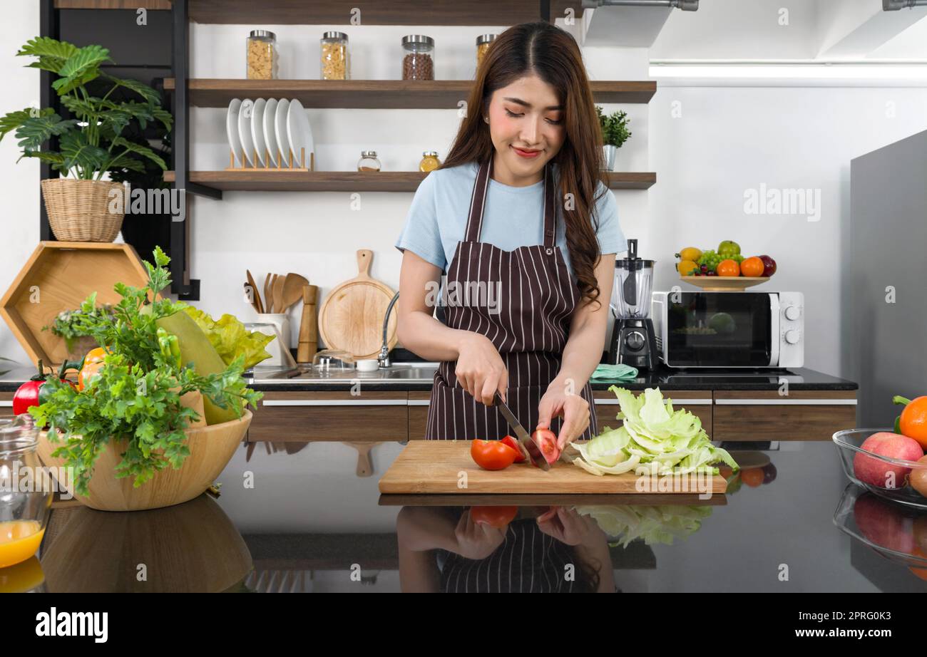 Jeune femme de ménage asiatique vêtue d'un tablier, tranche de tomate et laitue croquante sur une chop en bois. Le comptoir de cuisine plein de différents types de fruits et légumes. Banque D'Images