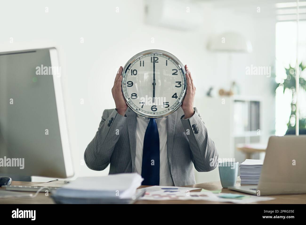 HES a eu du temps à penser : un homme d'affaires inidentifiable tenant une horloge devant son visage tout en étant assis à son bureau. Banque D'Images