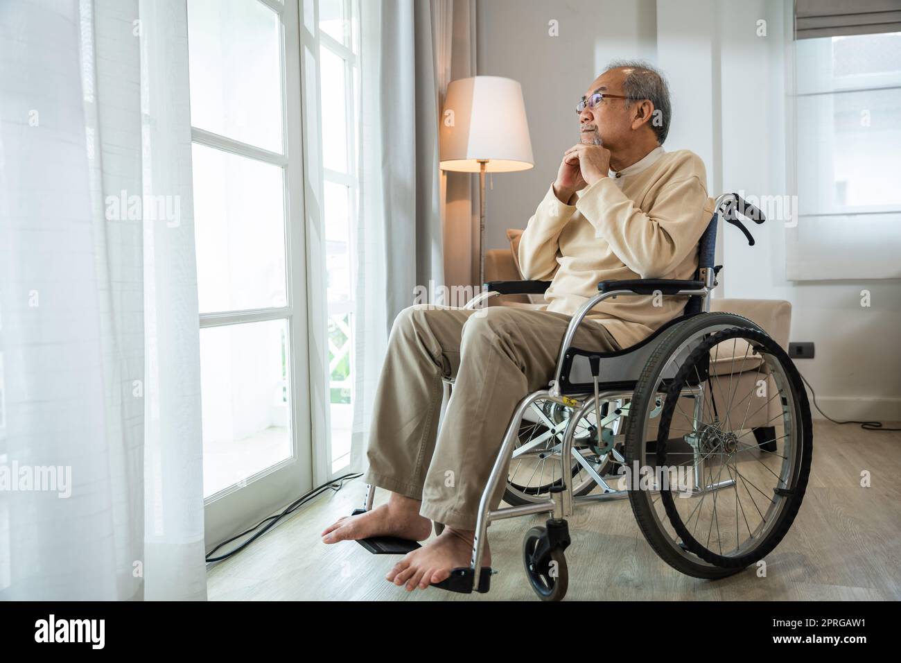 Homme asiatique âgé handicapé assis seul en fauteuil roulant et regardant par la fenêtre à l'hôpital Banque D'Images