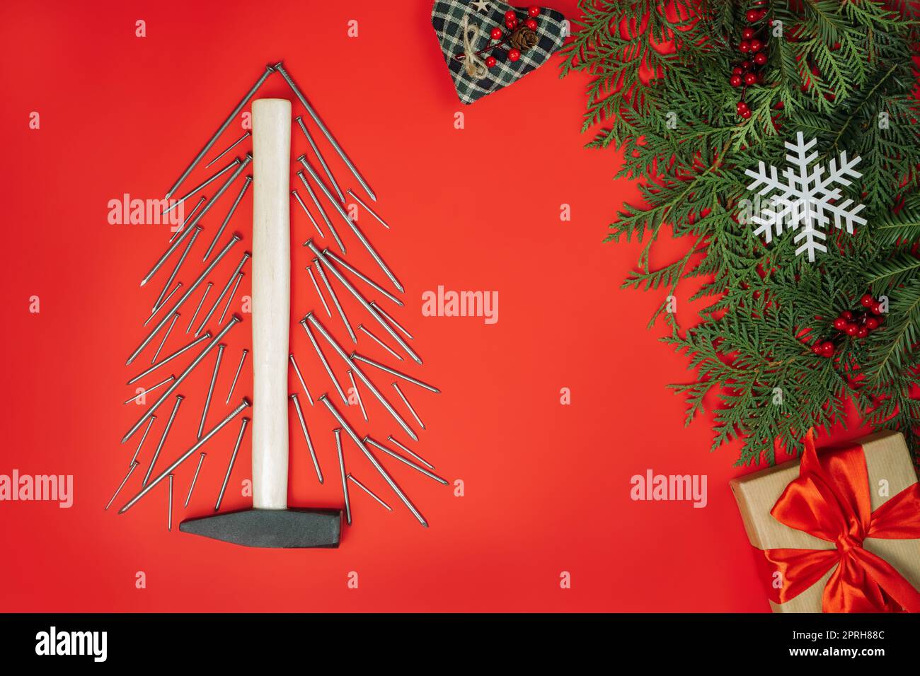 Clous longs et marteau, disposés en forme d'arbre de Noël avec branches d'épinette et boîte cadeau sur fond rouge.Pose à plat Banque D'Images