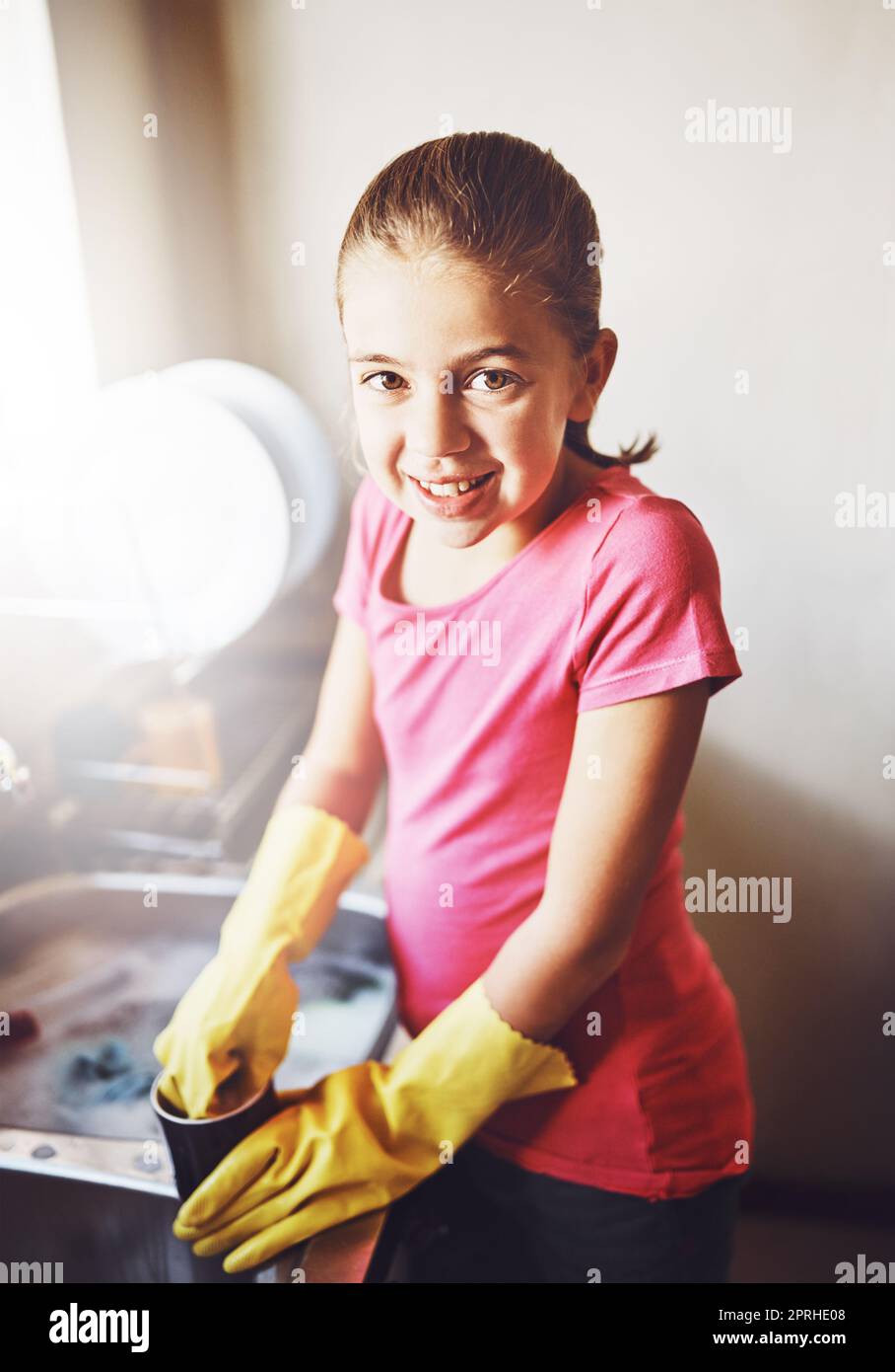 Aider maman avec des plats. Portrait d'une jeune fille gaie lavant des plats avec des gants jaunes à la maison tout en regardant l'appareil photo. Banque D'Images