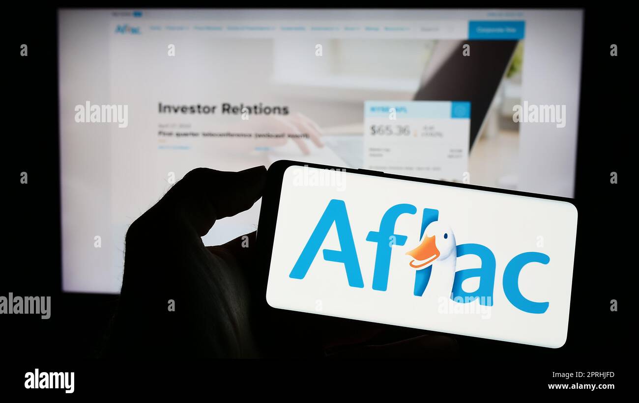 Personne tenant un téléphone cellulaire avec le logo de la compagnie d'assurance américaine Aflac Inc. À l'écran devant la page Web d'affaires. Mise au point sur l'affichage du téléphone. Banque D'Images