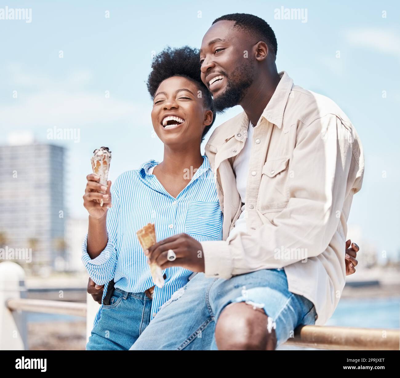 Couple heureux à la plage en train de manger un cône de glace pendant une journée dans la nature pendant les vacances de printemps. Homme noir et femme parlant et riant tout en embrassant et ayant le dessert en vacances en bord de mer. Banque D'Images