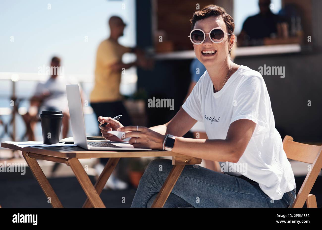 Je me contente de reprendre du travail. Portrait d'une jeune femme joyeuse travaillant sur son ordinateur portable tout en étant assise à une table près d'une promenade de plage Banque D'Images