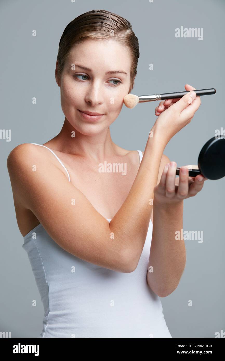 Création d'une œuvre d'art. Studio prise de vue d'une jeune femme attrayante appliquant un maquillage avec un pinceau sur fond gris. Banque D'Images