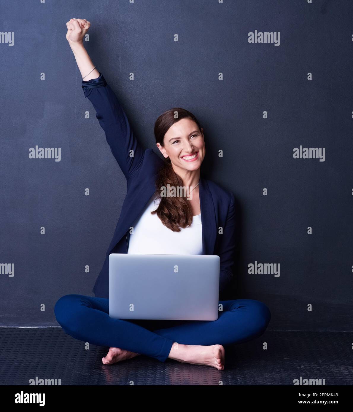 Une carrière et une passion se conjuguent. Une femme d'affaires excitée assise sur ses genoux avec son ordinateur portable sur un fond sombre. Banque D'Images