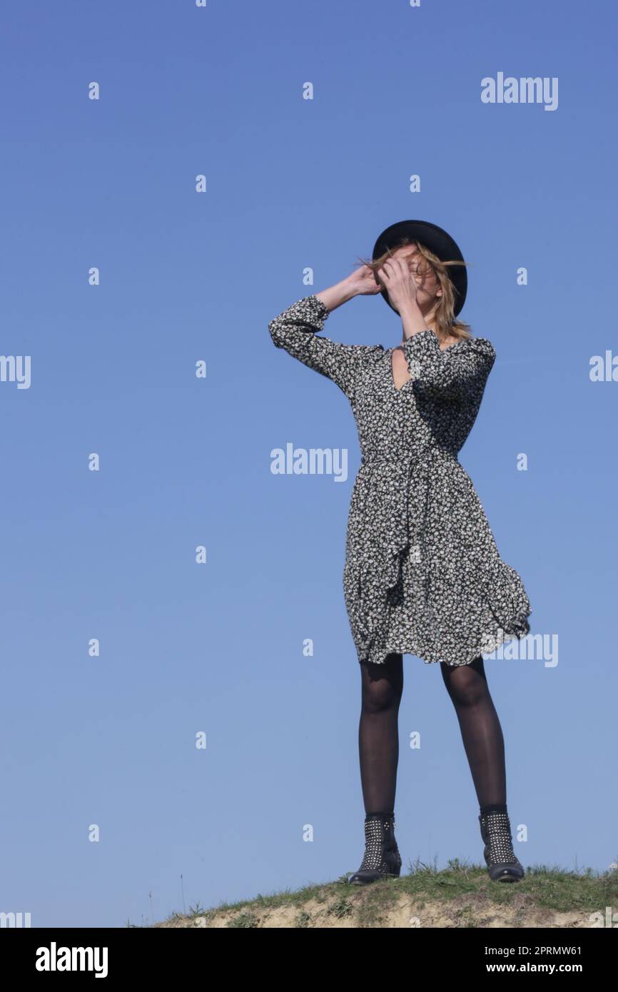 Série de photos de mannequin en robe fleurie boho viscose et chapeau avec ciel bleu clair derrière. Portrait de mode en extérieur avec lumière naturelle. Banque D'Images