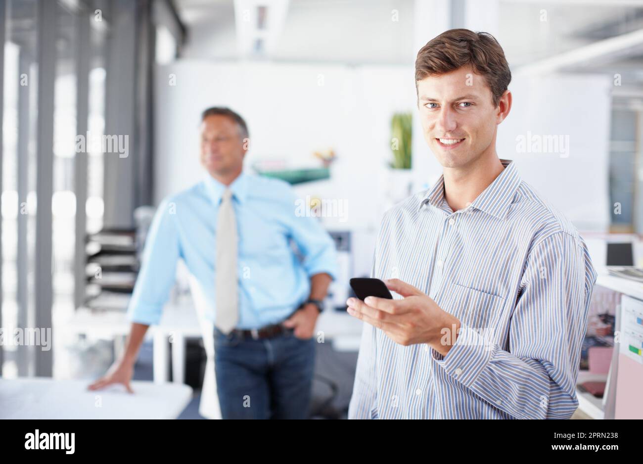 Utilisation des outils du métier. Un jeune professionnel tenant un smartphone tandis qu'un collègue se tient en arrière-plan. Banque D'Images