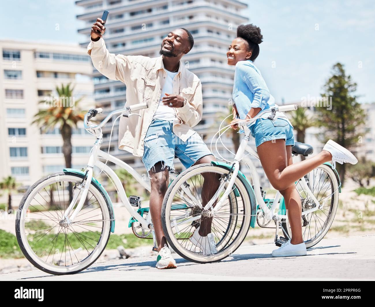 Selfie, vélo et rendez-vous avec un couple en vélo sur une promenade pendant l'été avec amour, romance et affection. Vélo, photographie et plaisir avec une femme et un homme noirs dehors et une ville en arrière-plan Banque D'Images