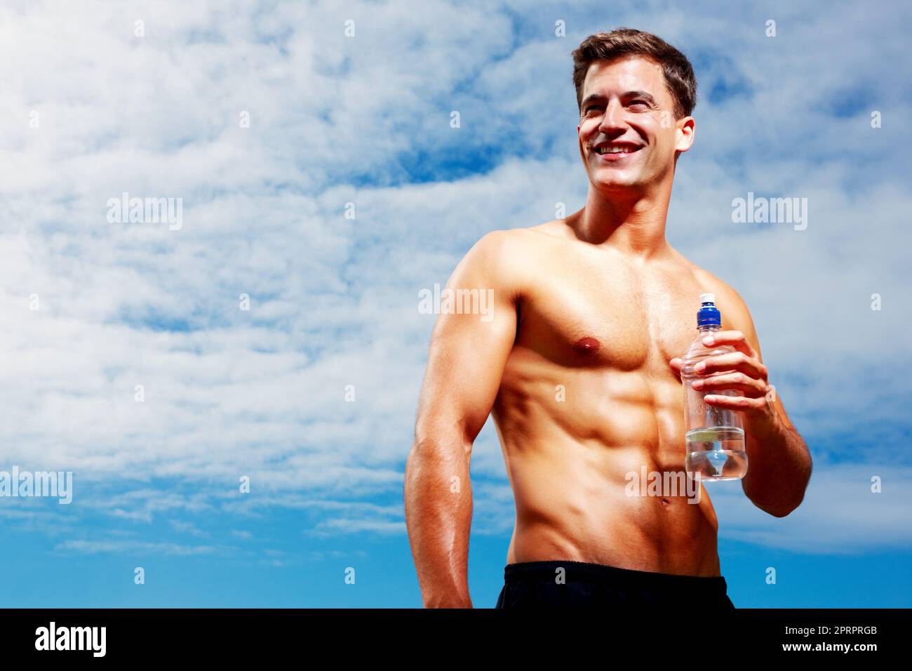 Un jeune homme heureux qui regarde loin avec une bouteille d'eau. Portrait d'un jeune homme heureux regardant loin tout en tenant une bouteille d'eau contre le ciel Banque D'Images