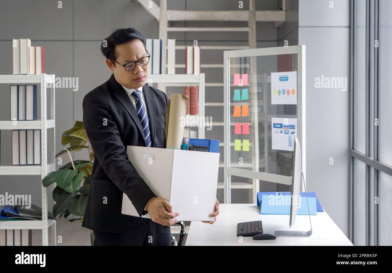 Le Manager asiatique en costume noir se sent triste, stressé et malheureux visage tenant une boîte en papier des effets recueillis au bureau le jour du feu. Idée pour les chômeurs, crise du travail. Banque D'Images