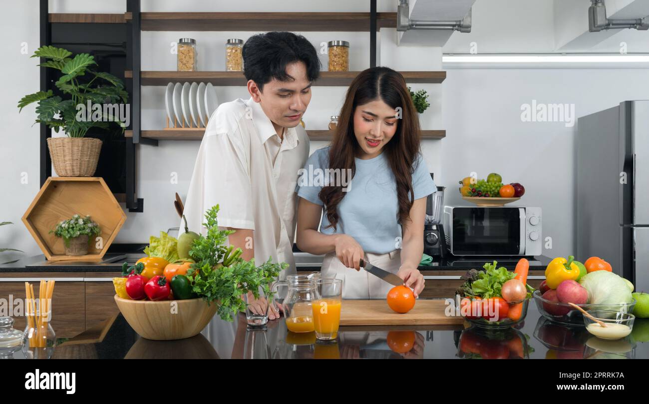 Un couple asiatique passe du temps ensemble dans la cuisine. Jeune femme coupant des fruits orange sur une planche à découper en bois tandis que son petit ami se trouve à côté. Divers fruits et légumes sont placés sur le comptoir. Banque D'Images