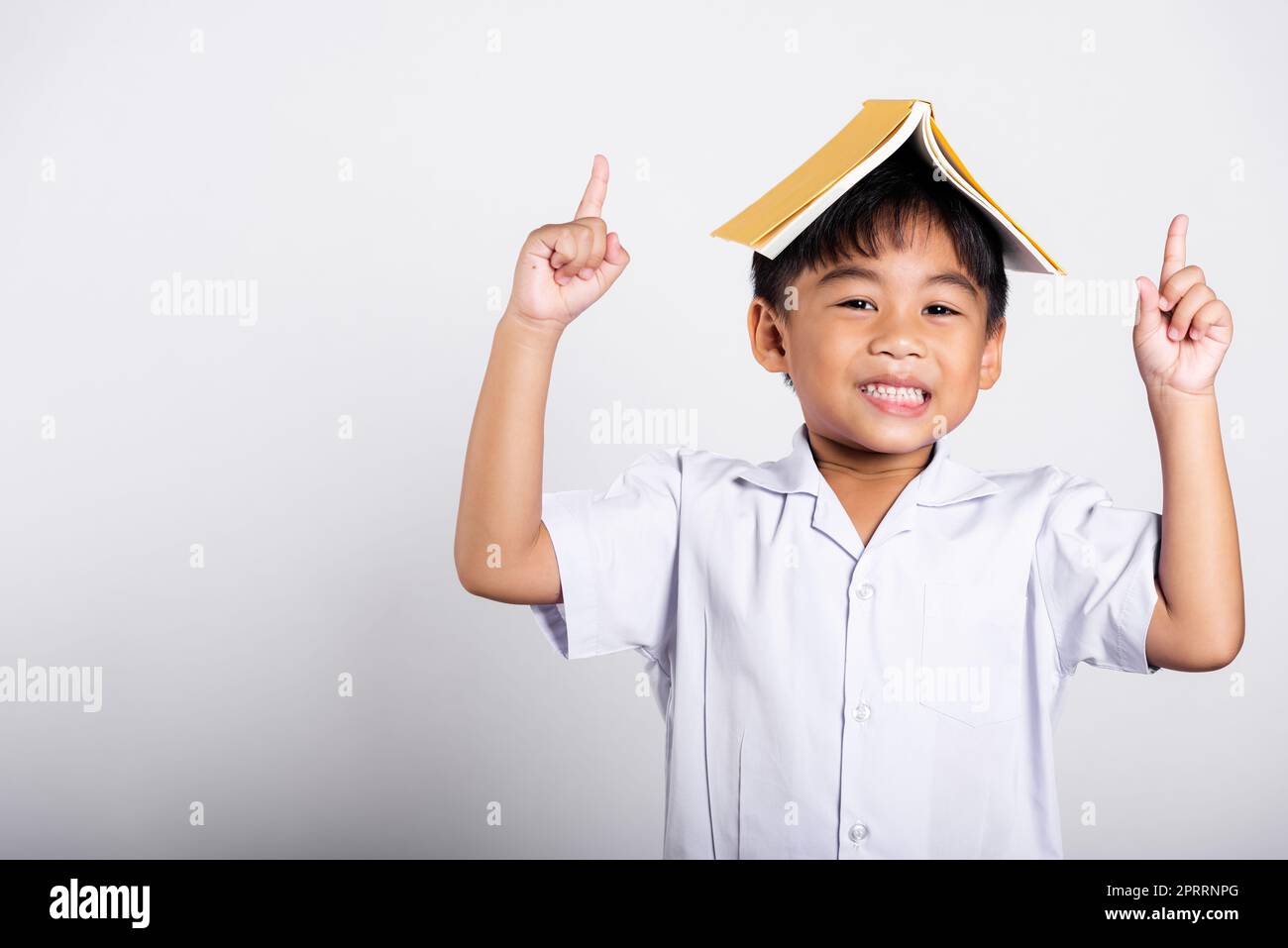 Asiatique adorable tout-petit souriant heureux portant étudiant thaïlandais uniforme pantalon rouge stand tenant le livre sur la tête comme le toit Banque D'Images