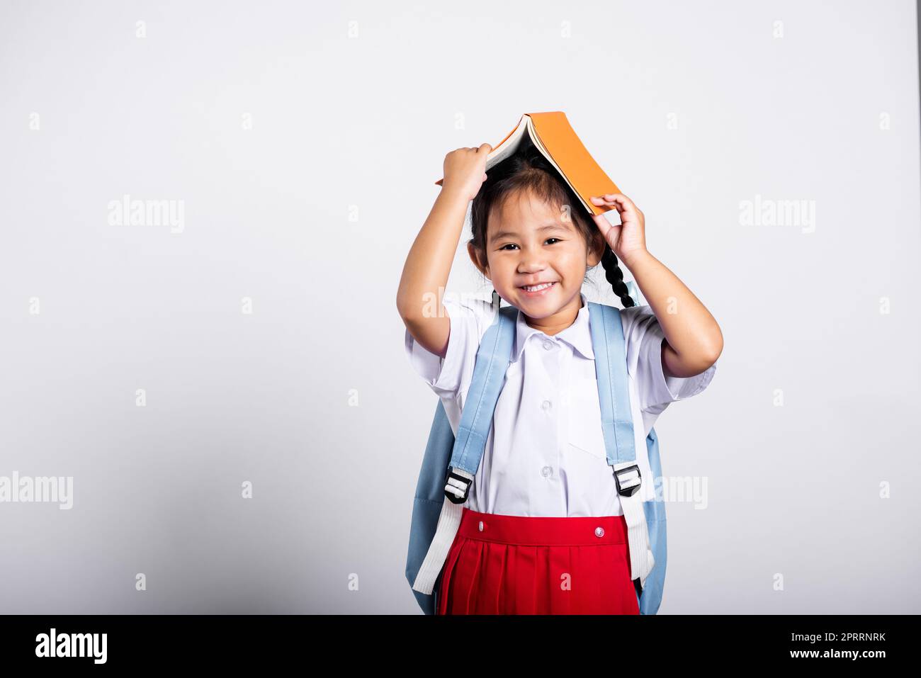 Asiatique adorable tout-petit souriant heureux portant étudiant thaïlandais uniforme rouge jupe stand tenant le livre sur la tête comme le toit Banque D'Images