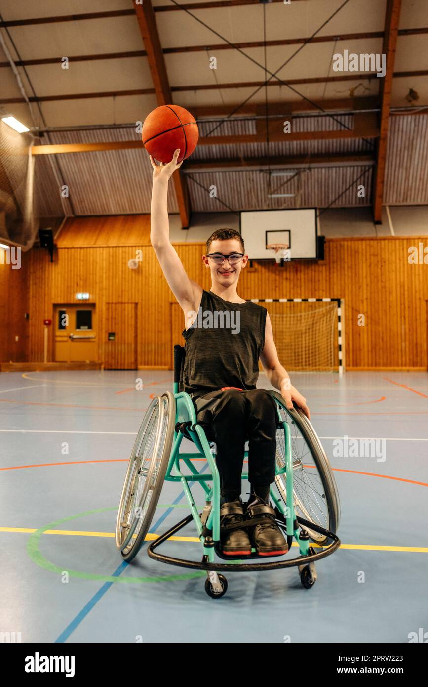 Fille souriante qui équilibre le basket-ball tout en étant assise en fauteuil roulant sur le terrain de sport Banque D'Images