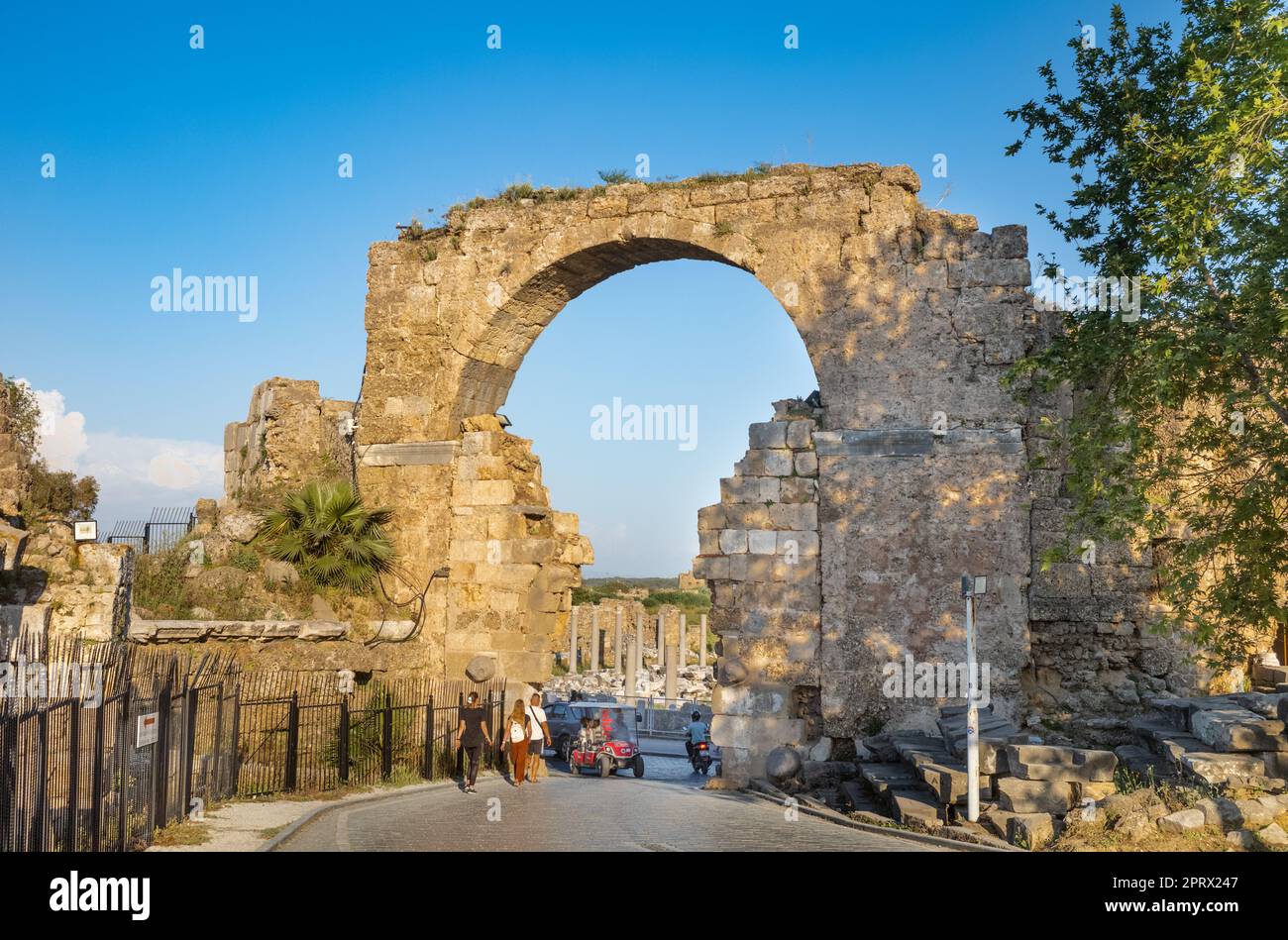 Les touristes se promènent à travers la porte Monumental près du monument Vespasien tandis qu'un buggy électrique se déplace l'autre côté de l'ancienne ville romaine en Turquie (T Banque D'Images Les touristes se promènent à travers la porte Monumental près du monument Vespasien tandis qu'un buggy électrique se déplace l'autre côté de l'ancienne ville romaine en Turquie (T Banque D'Images