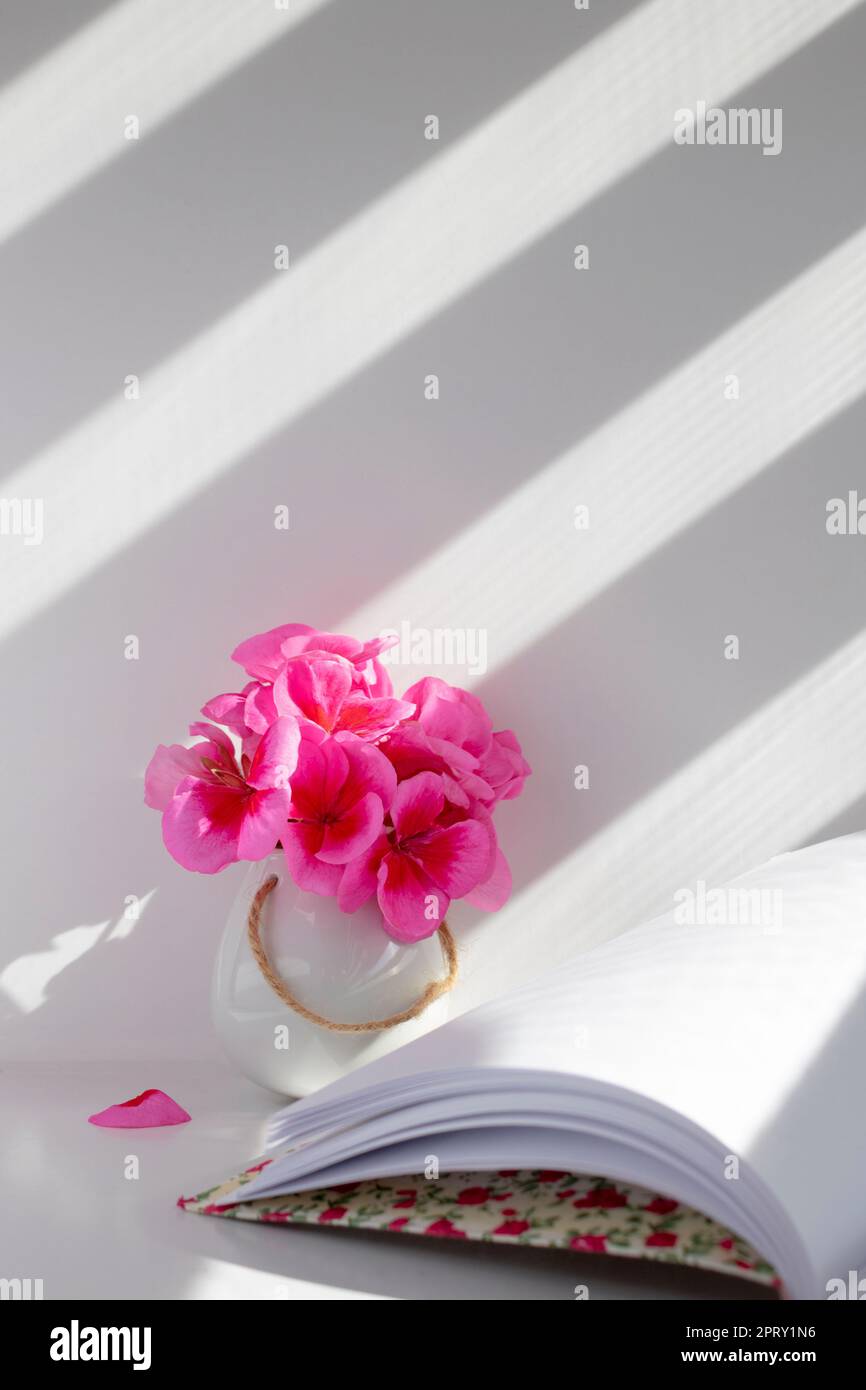 petit vase blanc avec géraniums roses et un livre ouvert avec des feuilles blanches Banque D'Images