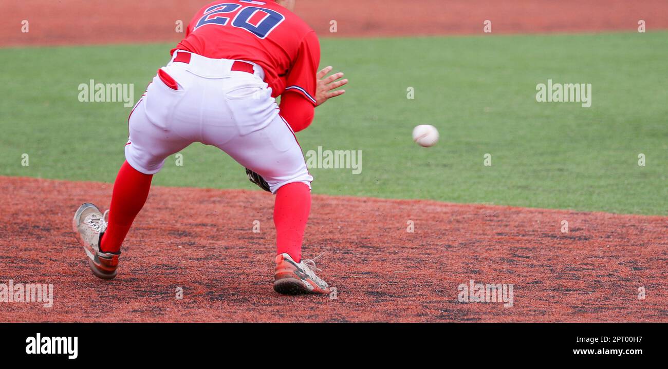 Vue arrière du troisième joueur de base qui joue un ballon lors d'un match de baseball d'école secondaire sur un terrain de gazon vert et rouge. Banque D'Images