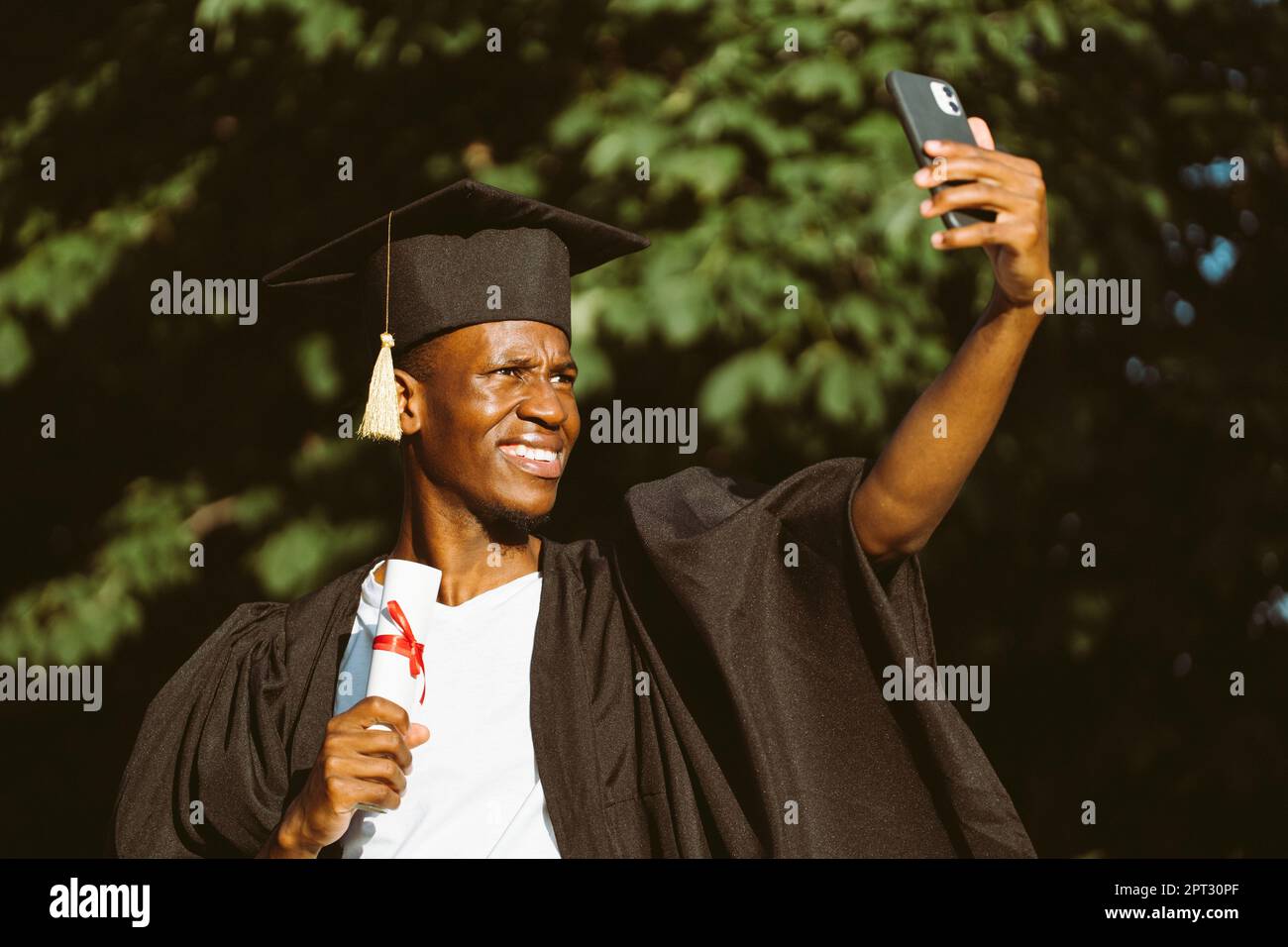 Joyeux homme noir diplômé de l'université prenant selfie avec l'appareil photo avant de smartphone avec diplôme d'enseignement supérieur. Étudiant en manteau noir et chapeau Banque D'Images