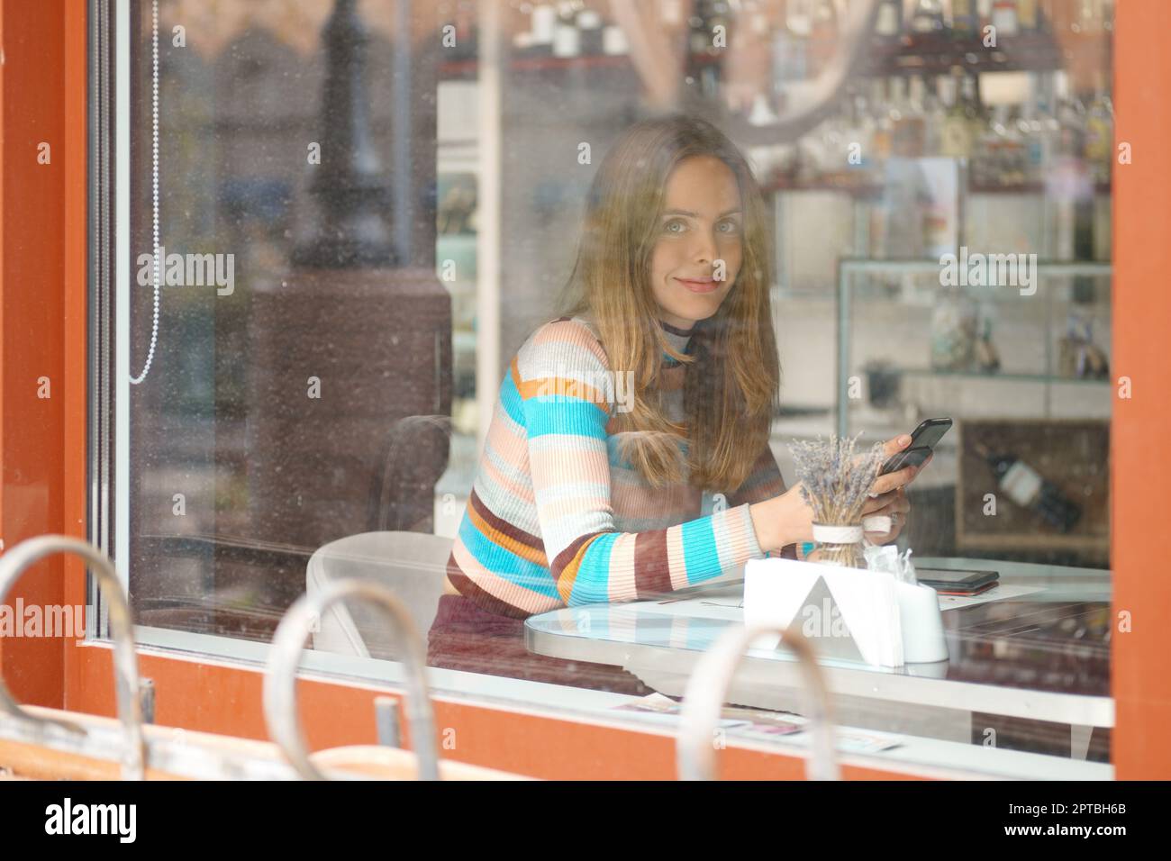Jolie fille aux cheveux longs, assise dans une caffe et bavardant sur un téléphone portable. Photo prise de l'extérieur, avec une vue à travers la fenêtre. Banque D'Images