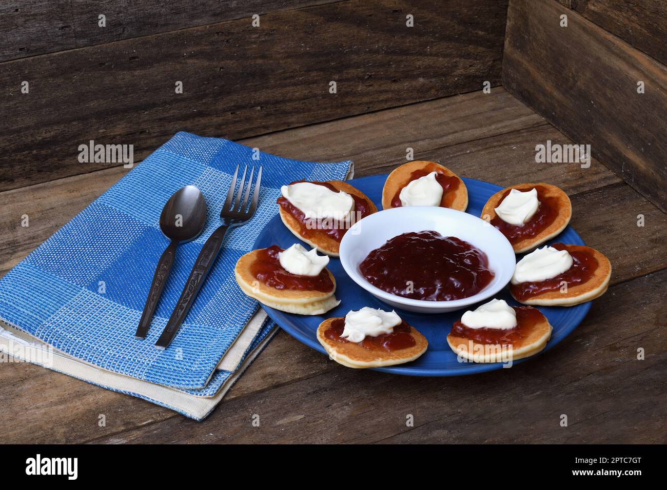 Pikelettes/crêpes/Battercakes avec confiture de fraise et crème fouettée sur une assiette bleue avec serviette, cuillère et fourchette dans un cadre rustique en bois Banque D'Images