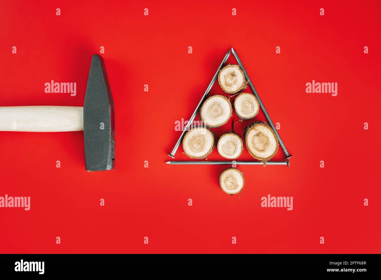 De longs clous et des coupes de bois ou des dalles de branches, disposés en forme d'arbre de Noël avec un marteau sur fond rouge.Pose à plat Banque D'Images