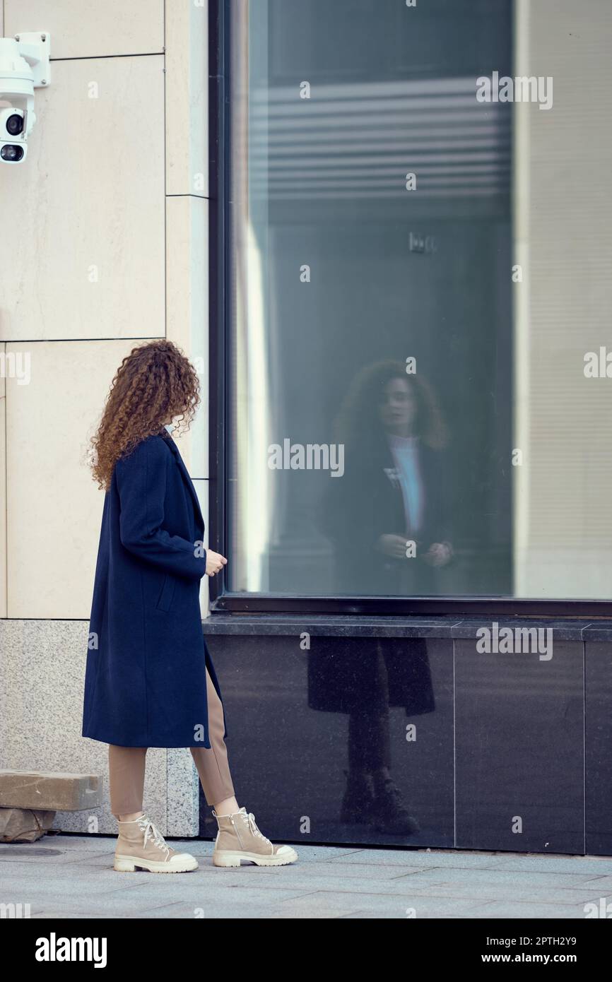 Une femme en manteau méconnaissable marche le long du trottoir et regarde son reflet dans les fenêtres Banque D'Images