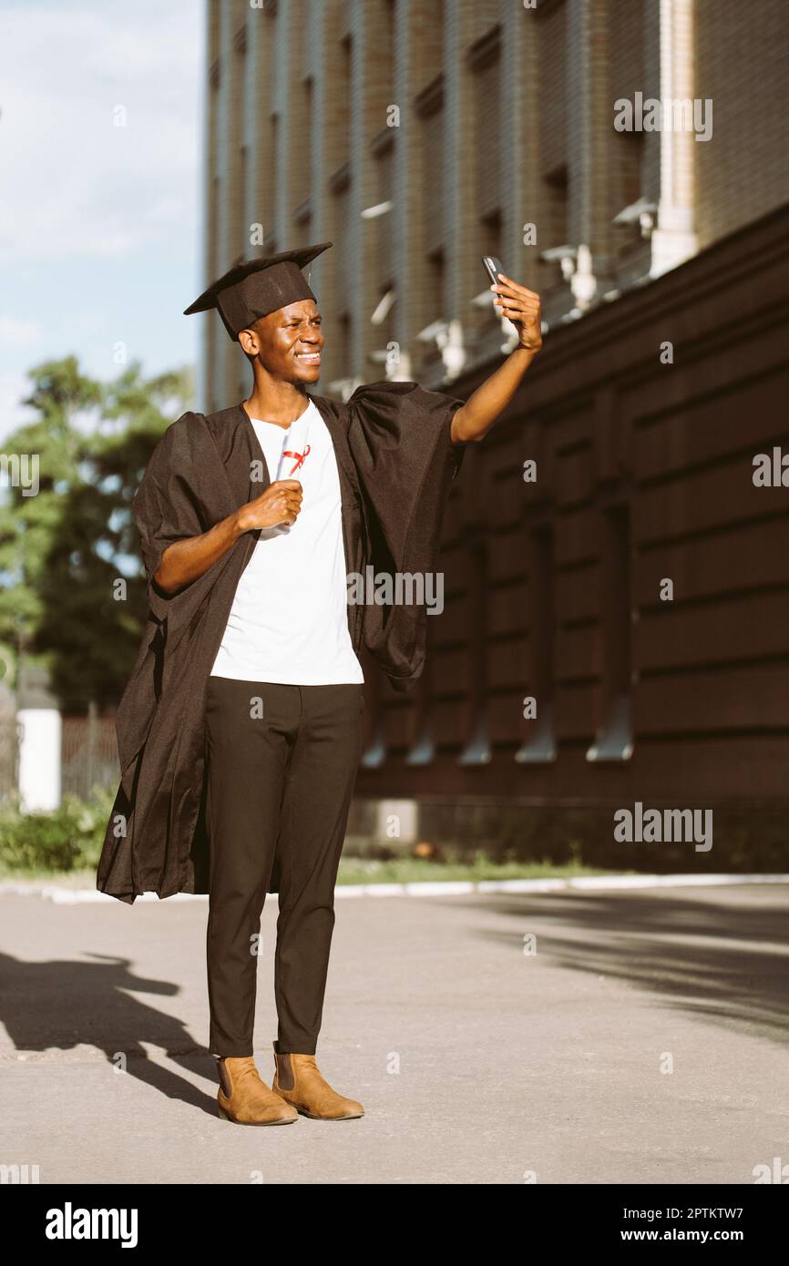 Un diplômé afro-américain souriant de l'université prend des photos avec l'appareil photo avant d'un smartphone avec un diplôme d'enseignement supérieur. Élève dans le manteau noir Banque D'Images